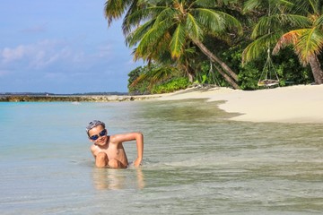 Happy little cute eight year old boy in swimming goggles is sitting in shallow seawater on exotic tropical white sandy beach with palm trees on background. Play,relaxing.Summer Leisure activities. 