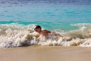 Laughing little cute boy in transparent splashes of rushing wave. Happy eight year old boy enjoys splashing waves in shallow sea water on exotic tropical beach.  Summer active leisure.Family vacation.