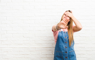 young blonde woman covering face with hand and putting other hand up front to stop camera, refusing...
