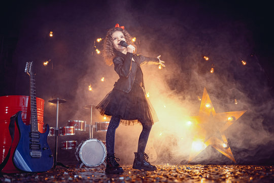 Beautiful girl with curly hair wearing leather jacket, boots sing into a wireless microphone for karaoke like rock star in recording studio or stage. Smoke on background.