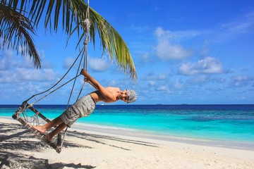 Happy little cute eight year old boy  is swinging on coconut palm tree seesaw on exotic tropical beach on background. Beach seesaw under palm trees. Play, relaxing.Summer Leisure activities. 