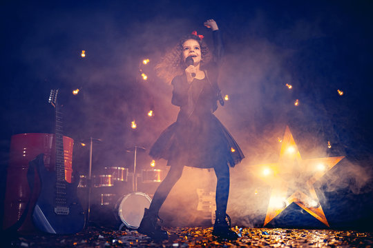 Beautiful girl with curly hair wearing leather jacket, boots sing into a wireless microphone for karaoke like rock star in recording studio or stage. Smoke on background.