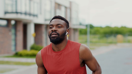 Active male runner in red sportswear and earphones jogging in the street. Outdoor portrait handsome african fitness guy performing cardio training for running marathon.