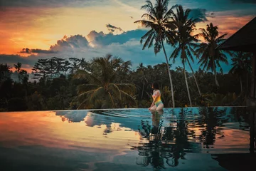 Wandcirkels aluminium Vrouw ontspant in een luxe overloopzwembad met uitzicht op de jungle bij zonsondergang in Ubud, Bali. Een meisje zit aan de rand van het overloopzwembad tegen de achtergrond van een heldere prachtige zonsondergang en de jungle. © Natallia