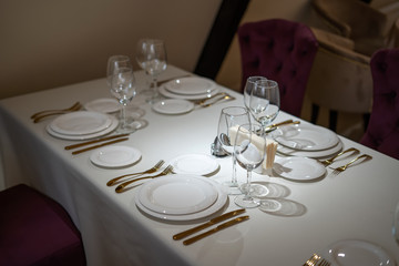 Chic and elegant, gold-plated cutlery and white plates, table setting with empty plates. Glasses in the light from the window. Side view.