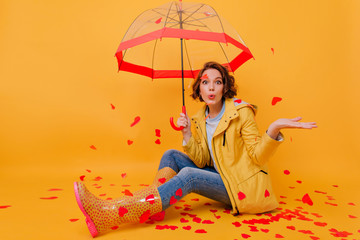 Indoor photo of spectacular girl wears rubber shoes and blue denim pants posing with umbrella. Portrait of joyful lady sitting on the floor with red paper hearts.