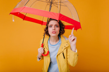 Beautiful girl with wavy hairstyle making funny faces while posing with trendy umbrella. Photo of unhappy white woman in autumn coat holding red parasol.