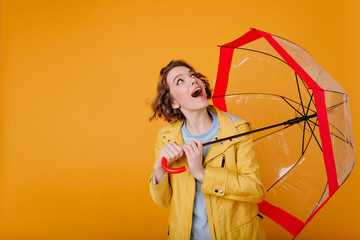 Surprised carefree girl in trendy coat looking up, holding umbrella. Studio shot of pretty woman with wavy hair posing with parasol on yellow background. © Look!