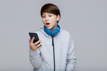 Portrait of young boy in hoodie with phone in hands. Gray background. Surprised youngster browsing internet on smartphone. Teenager in blue headphones listening music.