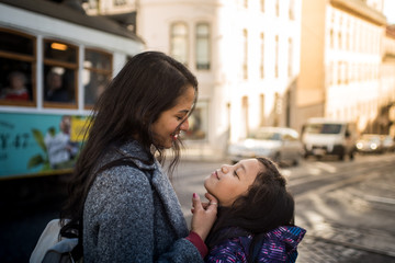 Latin mother and daughter looking affectionately outside on a Lisbon street, in the background there is a classic tram from Portugal