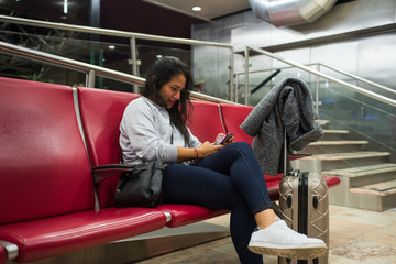 Young latina sitting in the airport terminal with her suitcase looking at the mobile phone. Latina chat with her cell phone while waiting for her flightv