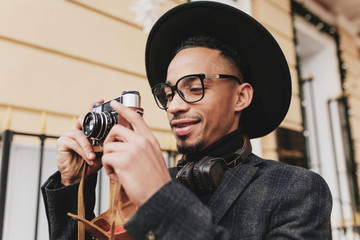 Cheerful male photographer with brown skin working outdoor in morning. Photo of positive african man wears dark clothes standing on the street with camera.