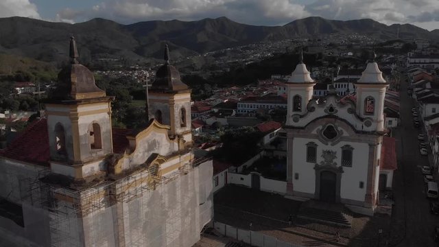 Aerial descend and tilt showing the two churches at central historical Gomes Freire square in the colonial mining town of Mariana in Minas Gerais, Brazil, with one in scaffolds as part of renovations
