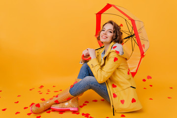 Fototapeta premium Relaxed pretty girl in yellow coat holding paper heart. Indoor photo of wonderful short-haired woman sitting under parasol during photoshoot in valentine's day.