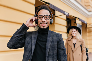 Elegant blonde girl in beige attire standing behind laughing african guy. Outdoor photo of chilling black man in headphones relaxing on the street.