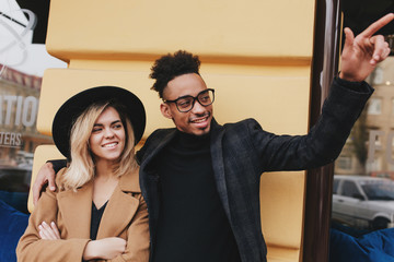 Smiling blonde girl in beige coat spending time with her male african friend. Good-looking black...