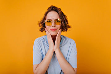Pale girl with light-brown hair touching her cheeks with pensive face expression. Studio shot of attractive trendy woman wears sunglasses.