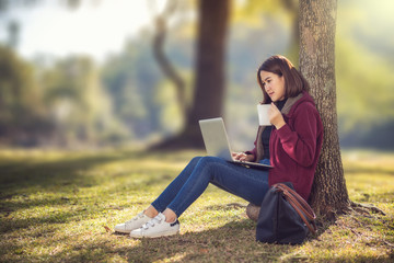 An Asian woman wearing a red coat in her hand, a white coffee cup sitting on the grass in a park working on a laptop. While sitting under a tree in the park with the strong sunlight from behind