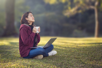 An Asian woman wearing a red coat in her hand, a white coffee cup sitting on the grass in a park working on a laptop. While sitting under a tree in the park with the strong sunlight from behind
