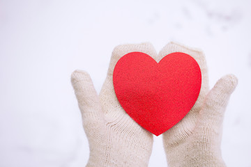 Valentines day background. Hands in white knitted mittens hold a red heart. Red Heart in hands. White snow background. Space for text.