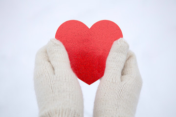 Valentines day background. Hands in white knitted mittens hold a red heart. Red Heart in hands. White snow background. Space for text.