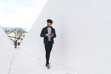 Trendy positive ethnic man using tablet while walking along urban street tunnel