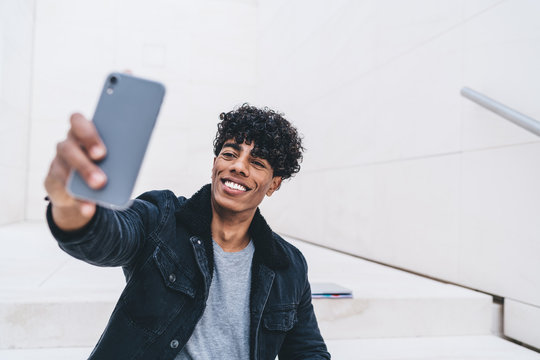 Smiling male blogger in stylish casual wear using modern smartphone camera for taking selfie on urban setting,positive hipster guy posing while making picture on cellular for sharing to virtual friend