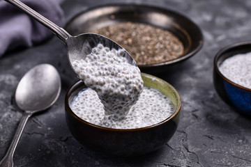 Spoon filled with chia seed pudding made with dry seeds soaked in milk in small bowls with dry seeds in background