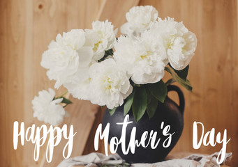 Happy Mother's day text handwritten on stylish peony bouquet in black  vase on rustic wooden background. White peonies rural still life. Happy Mothers day greeting card
