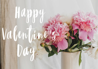 Happy Valentine's day text handwritten on lovely peony bouquet in sunny light on rustic window sill. Stylish pink and white peonies in vase on wooden background.