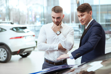 Mature salesman showing cars for sale at the dealership to his male client, copy space. Man buying new auto