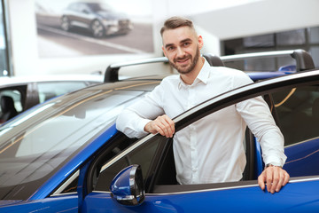 Happy handsome man smiling to the camera while examining new car on sale at dealership salon, copy space