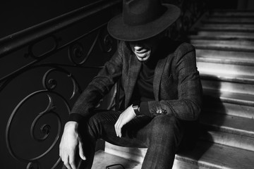 Black and white portrait of smiling man in elegant hat. Good-humoured african model sitting on stairs in the dark.