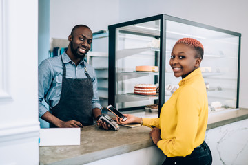 Ethnic pleased employee waiting while customer making mobile payment in cafe