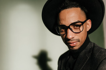 Fototapeta premium Close-up shot of interested guy in glasses posing in dark room. Portrait of black male model in trendy hat isolated on gray background.
