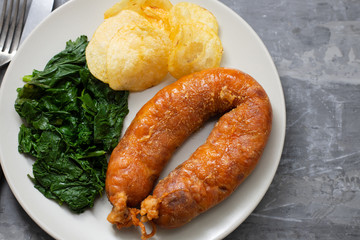 fried smoked sausage with potato chips and greens on white plate on ceramic background