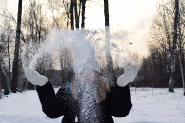 Young woman in a grey knitted beanie, blue scarf and black down jacket is playing with the snow in...