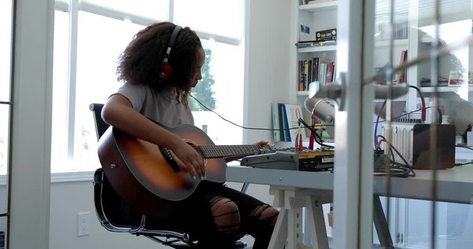 Teenage girl with headphones playing guitar, recording music in home office