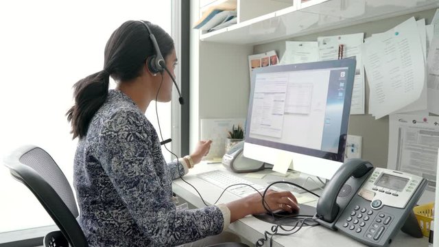 Female Clinic Office Manager With Headset Talking On Telephone At Computer In Office