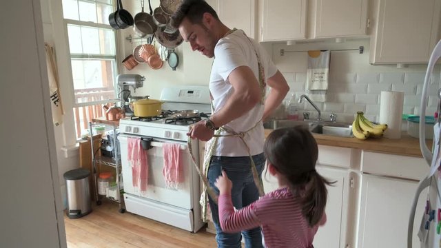 Daughter helping father tie apron in apartment kitchen