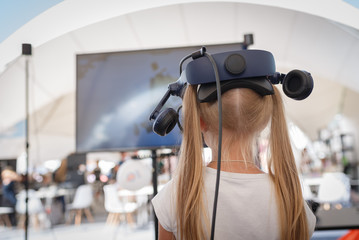 A little girl in a virtual reality helmet stands alone in front of a TV in an entertainment center