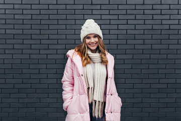 Stunning european girl in trendy knitted hat enjoying leisure time. Outdoor portrait of stylish white woman standing on urban background with shy smile.