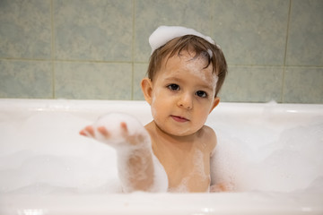 Cute baby girl taking a bath. looks at the camera and holds soap foam in hand. close up, soft focus, on the backgound is bathroom in blur