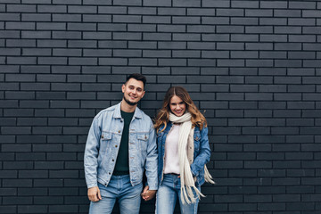 Excited girl in trendy denim outfit holding hands with boyfriend. Smiling loving couple standing together on bricked background.