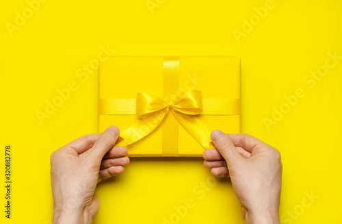 Male hands holding bright yellow gift present box with ribbon on yellow background top view. Flat lay holiday background. Birthday present, March 8, Mother's Day, Valentine's Day. Congratulation