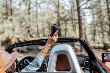 Woman enjoying traveling by a cabriolet in the forest, raising hands up with a coffee cup, view from the backside