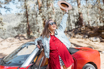 Portrait of a carefree woman enjoying road trip, standing with raised hands near the car in the forest. Carefree lifestyle and travel concept