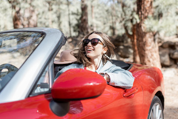 Portrait of a happy carefree woman driving convertible red car while traveling in the forest