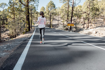 Young woman in sportswear jogging on the beautiful mountain road in the forest, wide view from the backside