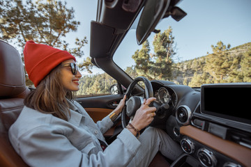 Happy woman dressed casually in jacket and red hat driving convertible car on the beautiful...